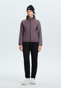 Giacca imbottita viola con zip e cappuccio, abbinata a pantaloni neri a gamba stretta e scarpe da ginnastica bianche. Il modello indossa un cerchietto nero.