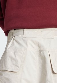 Svijetlosmeđi cargo hlače s teksturiranim materijalom, prilagođenim pojasom i malim izvezenim logom na leđa.