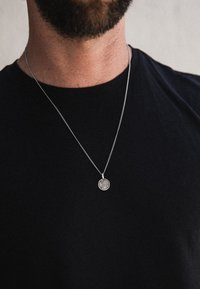Sølvvedhæng halskæde på en tynd kæde, med et cirkulært design og indgraverede detaljer, båret over en sort T-shirt med rund hals.