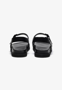 Svarte sandaler med en glatt skinnoverside, justerbar spenne, og flat gummisåle, med åpen bakdesign og minimalistisk stil.