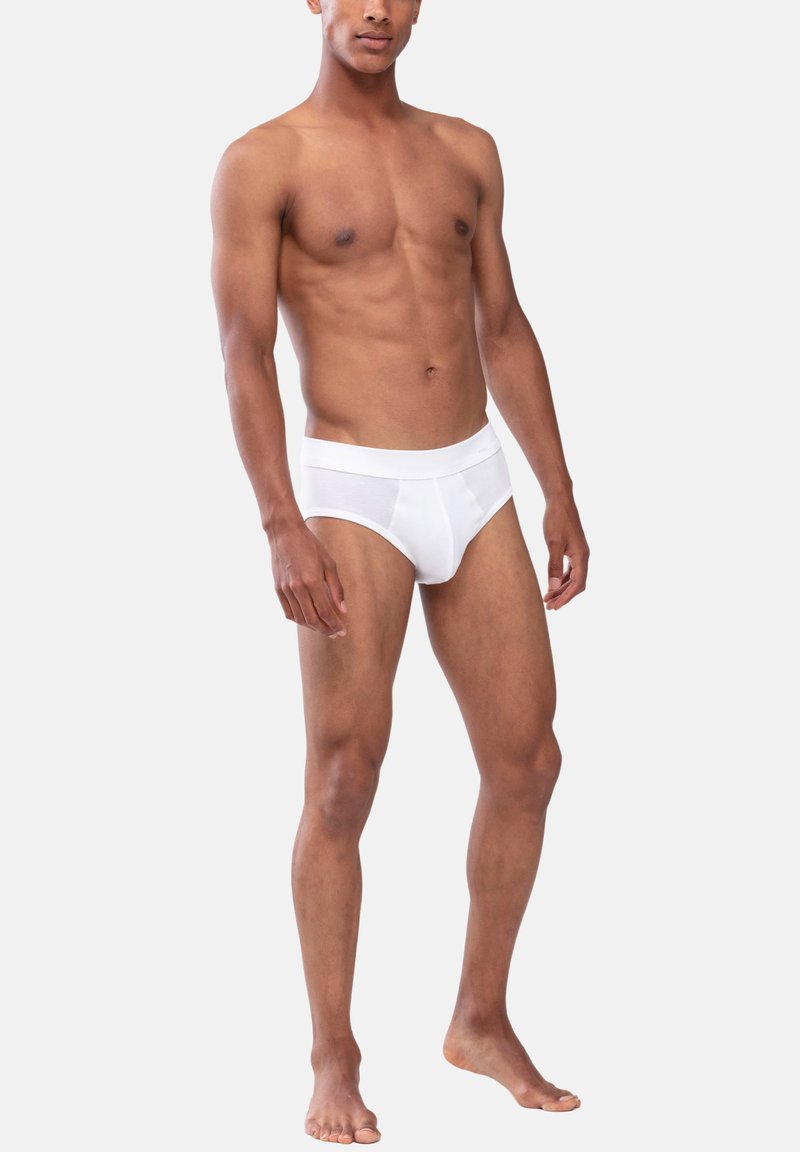 Modèle masculin debout pieds nus, ne portant que des slips blancs, sur un fond gris clair uni, montrant son physique et le style de sous-vêtements.