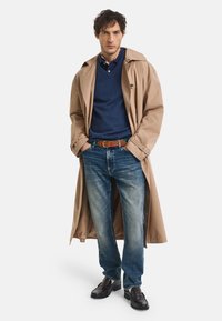 Beige Trenchcoat, marineblaues Sweatshirt, blaue Jeans, brauner Gürtel, dunkle Schuhe. Verfügt über eine lockere Passform und kontrastierende Texturen in den Schichten.