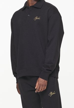 Black long-sleeve polo made of cotton with a ribbed hem. Features a gold embroidered logo on the chest and a classic collar design.