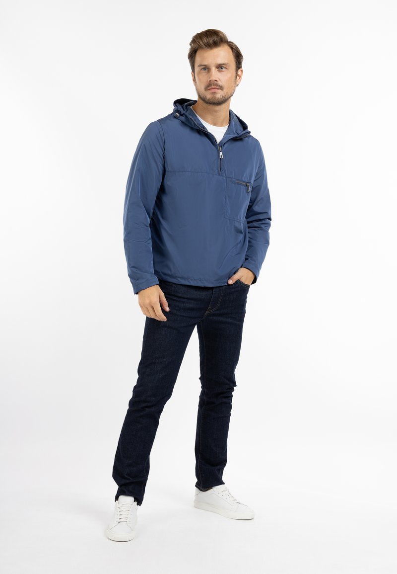 Veste légère à capuche bleue avec une fermeture éclair demi-longueur et une poche frontale, associée à un jean foncé et des baskets blanches sur un fond uni.