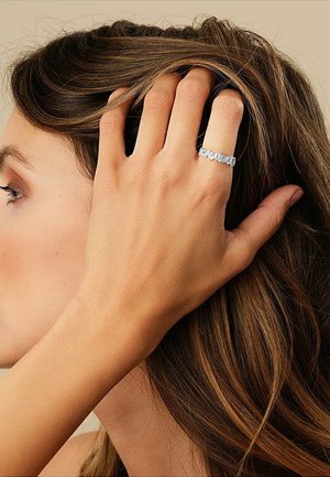 Silberner Ring mit mehreren klaren Steinen, die entlang des Rings angeordnet sind, getragen an einer Hand mit neutralem Hautton, vor einem sanften, gedämpften Hintergrund.