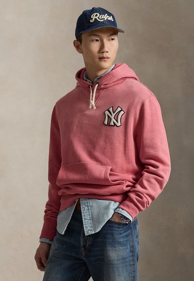 Sudadera con capucha de algodón rosa con bolsillo tipo canguro y logo bordado de NY, combinada con una camisa vaquera azul clara y unos vaqueros oscuros.
