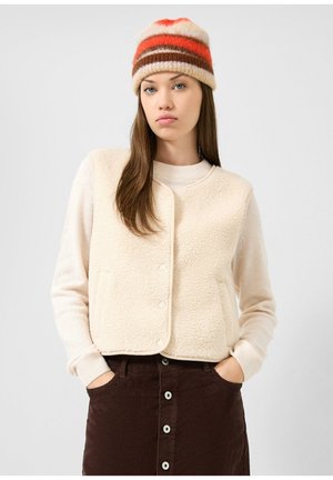 Femme portant une veste polaire crème, un pull beige, une jupe en velours côtelé marron avec boutons, et un bonnet tricoté avec des rayures orange, marron et crème.