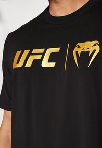 Sort t-shirt med et skinnende guld "UFC" logo og et gyldent slangemotiv. Stoffet ser glat og let ud med en crew neck design.