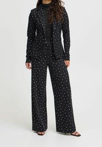 Kvinna klädd i en svart kostym med små vita hjärtan, svart polotröja och svarta öppna klackar, stående mot en enfärgad bakgrund.