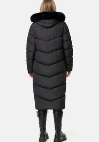Cappotto lungo nero in stile piumino con cuciture a zig-zag orizzontali, texture imbottita e cappuccio foderato in pelliccia sintetica. Indossato con stivali neri fino al ginocchio.