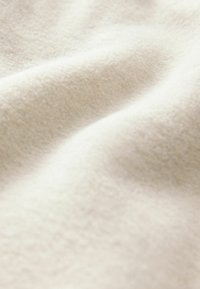 Kreeminsävyinen kangas, jossa on pehmeä ja tekstuuroitu pinta, joka näyttää lempeitä taitoksia ja ryppyjä. Materiaali vaikuttaa ylelliseltä ja lämpimältä.