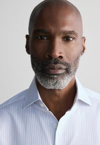 Chemise boutonnée à rayures bleu clair avec un col, portée par un homme avec une courte barbe grisonnante et une tête lisse et chauve. Fond uni.