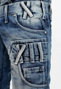 Pantalons cargo en denim bleu délavé avec plusieurs poches, coutures contrastées et boucles décoratives. Tissu texturé avec un aspect usé.