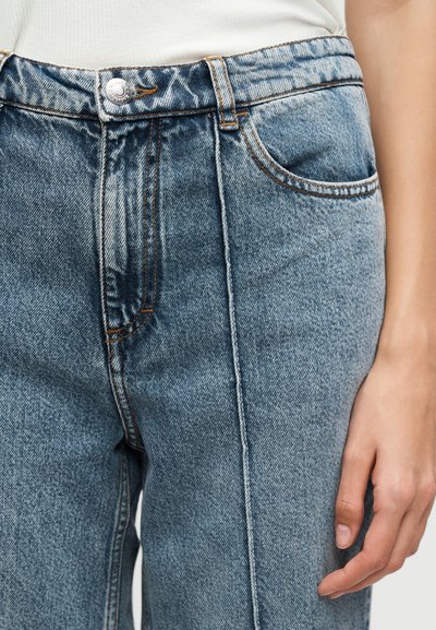 Κοντινό πλάνο μεσαίου μπλε τζιν τζιν με ορατό μπροστινό κουμπί, φερμουάρ και ένα χέρι που ακουμπά δίπλα στο δεξί μηρό.