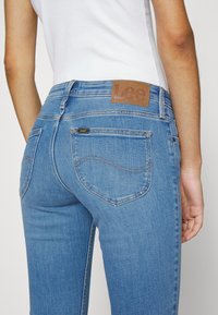 Blå denimjeans med en figursydd silhuett, brunt lädermärke vid midjan, accentuerad sömnad på fickorna och klassisk design på bakfickorna.