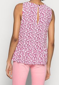 Top senza maniche con un motivo geometrico rosa e bianco, dettaglio a chiave sul retro e silhouette a peplum. Il tessuto appare liscio e leggero.