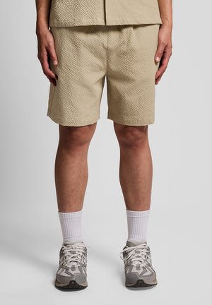 Uomo che indossa pantaloncini beige con texture, calzini bianchi alla caviglia e scarpe da ginnastica grigie e bianche, in piedi davanti a uno sfondo grigio uniforme.