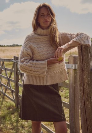 Femme en pull torsadé beige et jupe foncée s'appuyant sur une porte en bois à l'extérieur, tenant une pomme verte avec des champs en arrière-plan.