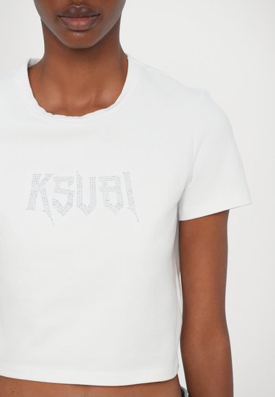 Balts īsām piedurknēm korsetes tops ar ribota apakšmalu un rhinestone akmeņiem, kas veido vārdu "KSVAL" priekšpusē. Mīksta kokvilnas materiāls, gluda tekstūra.
