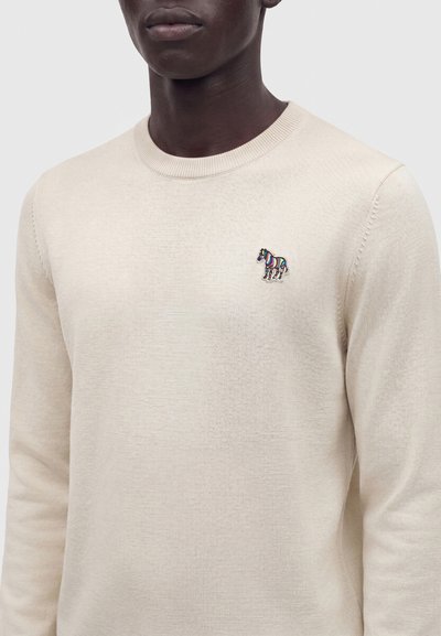 Homme portant un pull en maille beige clair à manches longues avec un petit logo zèbre multicolore sur la poitrine gauche.