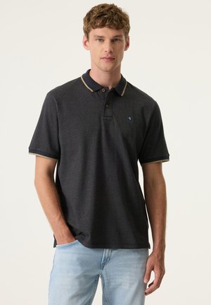Jonge man die een zwart poloshirt met korte mouwen draagt met beige rand en lichtblauwe spijkerbroek, staand met één hand in de zak.