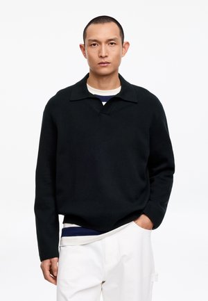 Homme portant un polo à manches longues noir par-dessus une chemise rayée bleu marine et blanc, avec un pantalon blanc, se tenant devant un fond blanc uni.