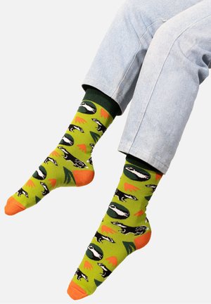 Zielone skarpetki z wzorem borsuków, pomarańczowymi palcami i zielonymi ściągaczami. Wzór zawiera drzewa i pomarańczowe akcenty na teksturowanym tle.