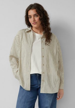 Vrouw met krullend haar, gekleed in een crème- en zwarte verticale gestreepte blouse over een wit shirt en blauwe spijkerbroek, staand tegen een lichte, effen achtergrond.