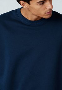 Tums zilas krēklis, kas izgatavots no mīksta auduma, ar ribotu apkakli un gludu tekstūru, ar minimāli redzamām detaļām.