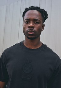 Svart bomullströja med präglad logotyp och två stjärnor på framsidan, med rund hals och korta ärmar, mot en ljus bakgrund.