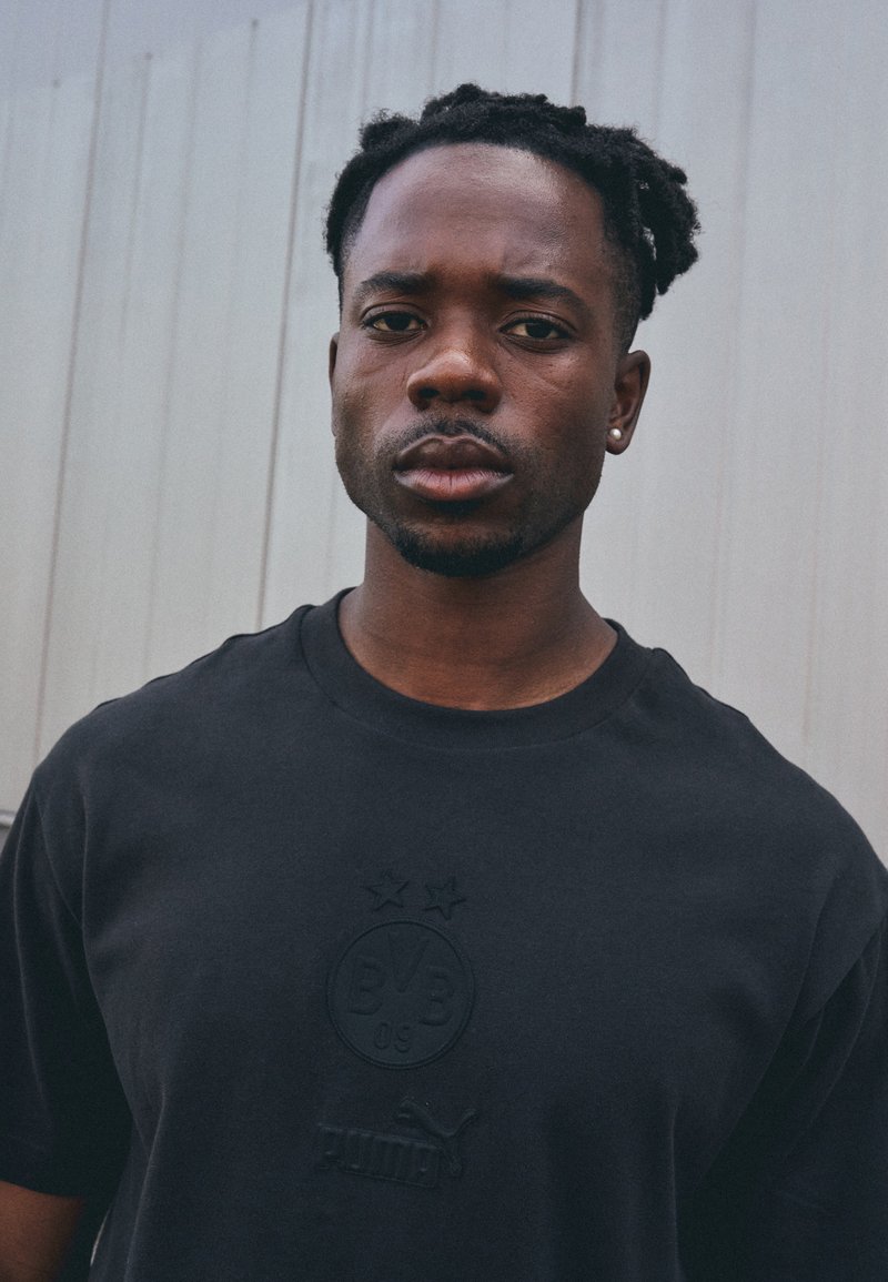 Svart bomullströja med präglad logotyp och två stjärnor på framsidan, med rund hals och korta ärmar, mot en ljus bakgrund.