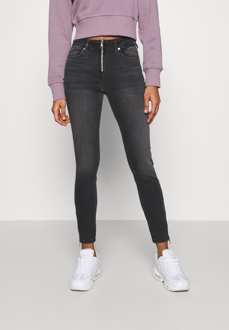 Persona vistiendo jeans skinny de tiro alto en negro desgastado con cremallera frontal y zapatillas blancas, sudadera corta de color lila visible a la altura de la cintura.