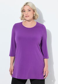 Fialová tunika s dlouhým rukávem a kulatým výstřihem, vyrobená z hladkého materiálu, s volným střihem a rovnými lemy. Doplňky jsou minimální.