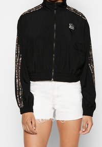 Svart kort jacka i lätt material, med elastiska ärmslut, dragkedja och mönstrade sidostråk. Adidas-logga på framsidan.