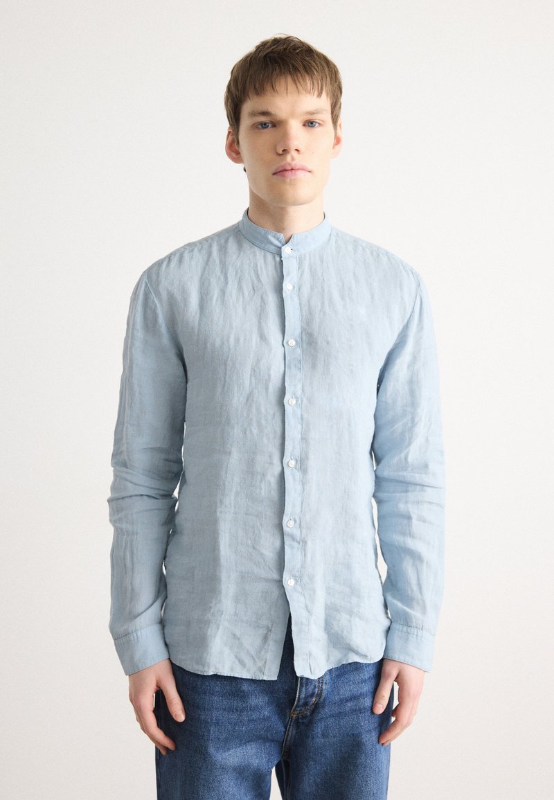 Himmelblaues, langärmeliges Leinenhemd mit Stehkragen, versehen mit einer Knopfleiste vorne und leicht zerknitterter Textur, kombiniert mit dunklen Jeans.