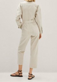 Lichtbeige jumpsuit met lange mouwen, een ceintuur in de taille en cropped pijpen. Textuurstof en twee achterzakken. Draagt met zwarte gespte sandalen.