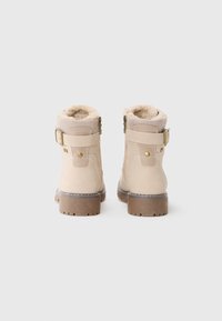 Botas de tobillo beige de ante con un acabado texturizado. Cuentan con un interior acogedor, detalles de correa y hebilla, y una suela de goma.