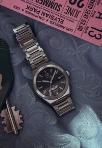 Timex-Armbanduhr aus Edelstahl mit schwarzem Ziffernblatt, Datumsanzeige, neben einem vintage Schlüssel und einem pinken Elvis-Konzertticket.