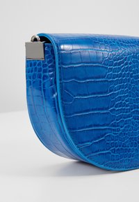 Borsa a spalla in finta pelle di cocodrillo blu con forma arrotondata, texture lucida e chiusura in metallo argentato. Presenta accenti in contrasto turchesi.
