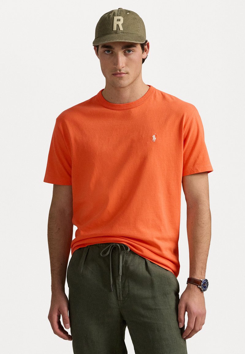 Orange bomull t-shirt med rund halsringning och korta ärmar, med en liten vit broderad logotyp på vänster bröst.