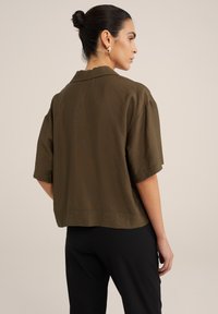 Maglietta verde oliva, taglio corto, realizzata con un tessuto morbido e texturizzato, con colletto piatto e maniche corte. Indossata con pantaloni neri.
