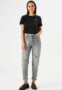 T-shirt en coton noir avec un design brodé, associé à un jean en denim gris taille haute et déchiré, et des chaussures noires à enfiler avec des accents dorés.