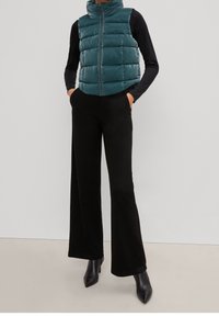 Veste matelassée turquoise à col montant, fermeture éclair et texture brillante, associée à un pantalon large noir et des talons pointus noirs.