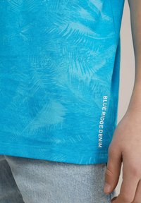 Mėlynos spalvos marškiniai trumpomis rankovėmis, dekoruoti palmių lapų raštu, šoninėje dalyje baltai išspausdinta "BLUE RIDGE DENIM".