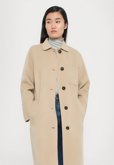 Beige long coat featuring a buttoned front, pointed collar, single pocket, and a soft, textured fabric. Worn over a gray turtleneck.