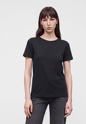 Jeune femme aux longs cheveux bruns portant un t-shirt noir uni à manches courtes et un jean foncé, debout devant un fond gris clair.