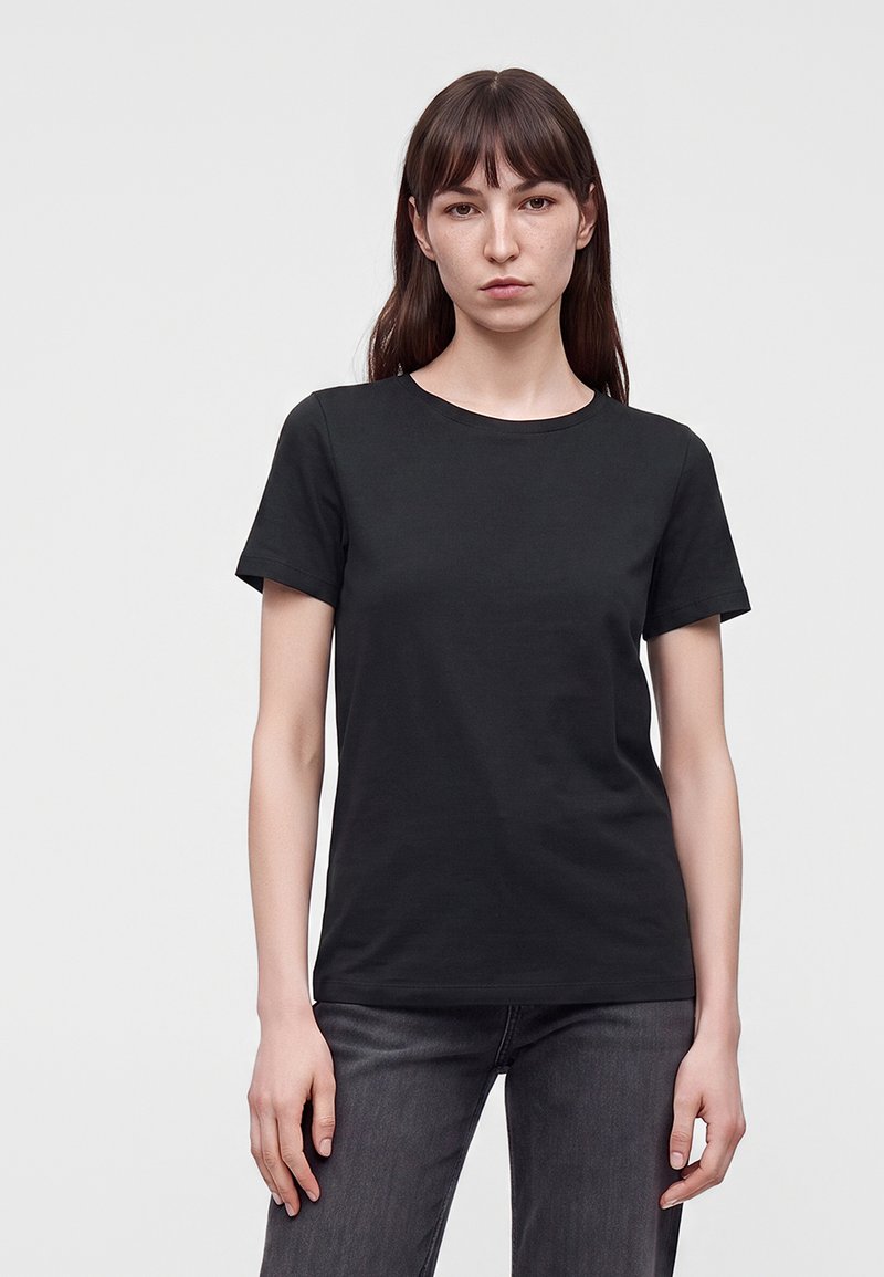 Jeune femme aux longs cheveux bruns portant un t-shirt noir uni à manches courtes et un jean foncé, debout devant un fond gris clair.