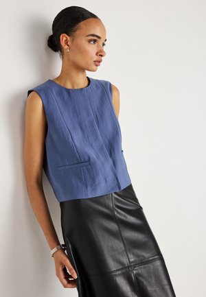 Frau mit hochgestecktem Haar trägt ein ärmelloses blaues Oberteil und einen schwarzen Lederminirock, sie steht seitlich an einer schlichten weißen Wand.