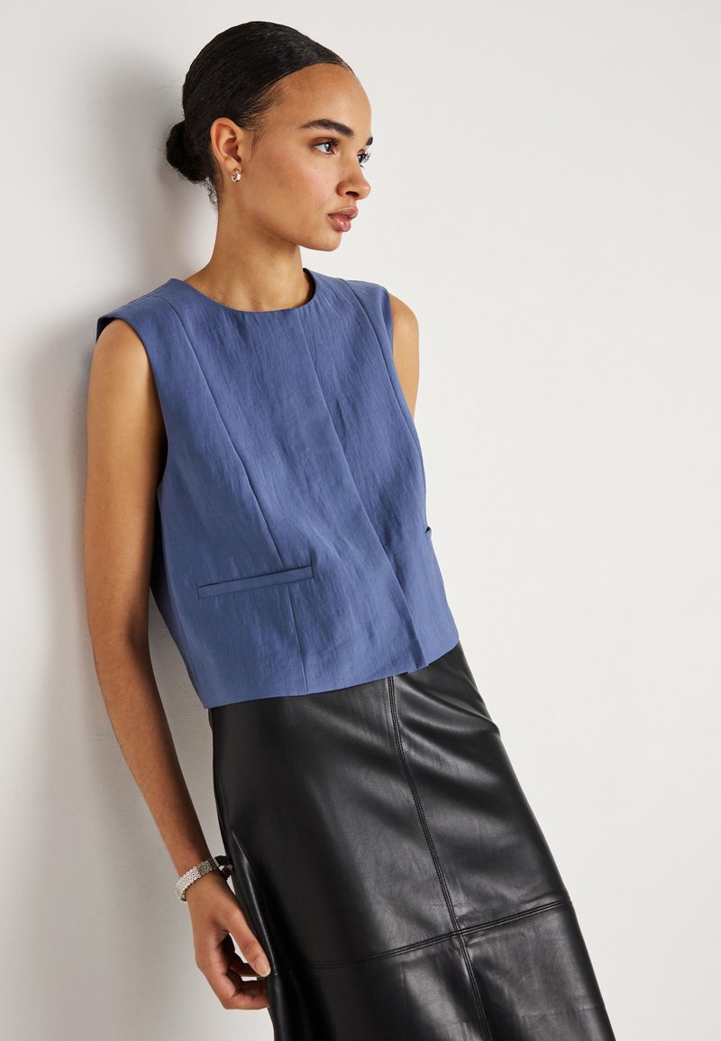 Γυναίκα με τα μαλλιά της σε κότσο φοράει μπλε αμάνικη μπλούζα και μαύρη δερμάτινη φούστα, στέκεται πλάγια μπροστά σε ένα απλό λευκό τοίχο.