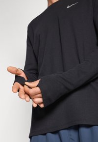 Chemise noire à manches longues avec texture côtelée et passe-pouces aux poignets. Présente un petit logo Nike gris sur la poitrine.